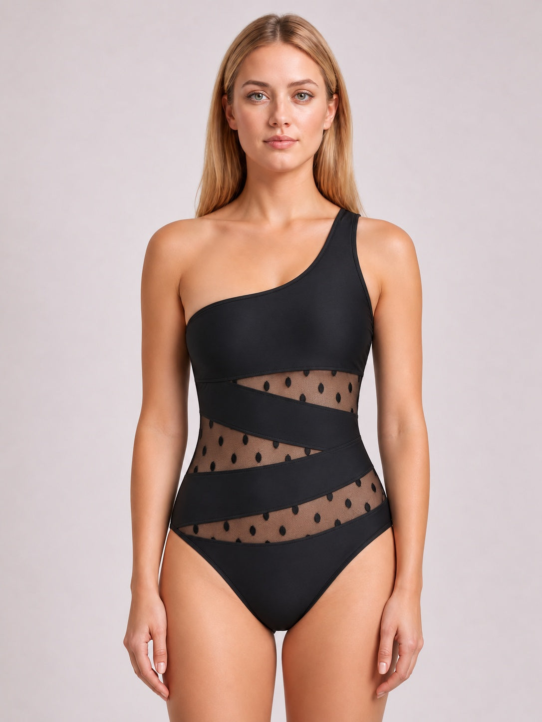 Front-facing model wears a black sleeveless one-piece swimsuit with an asymmetrical one-shoulder neckline, sheer dotted mesh panels, and crisscross waist bands.