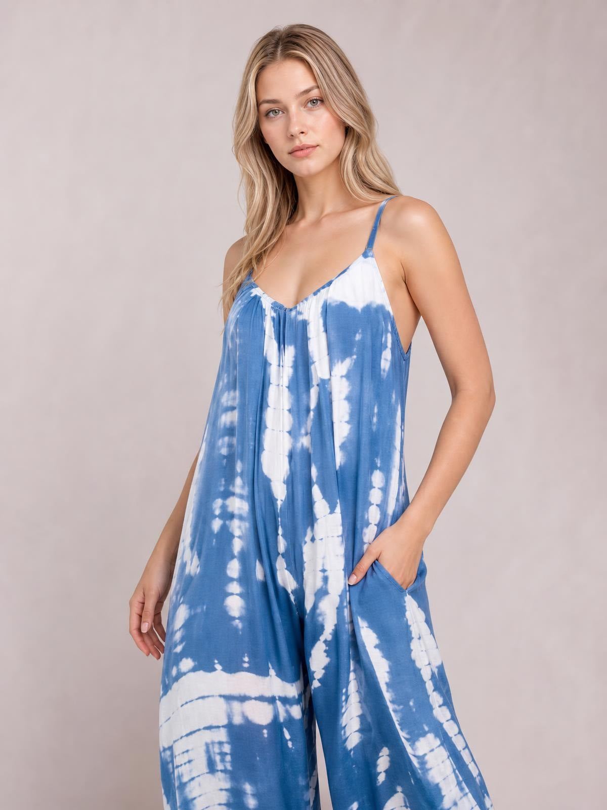 A model poses in a blue-and-white tie-dye V-neck spaghetti strap jumpsuit with a relaxed wide-leg silhouette and one hand tucked into a side pocket.