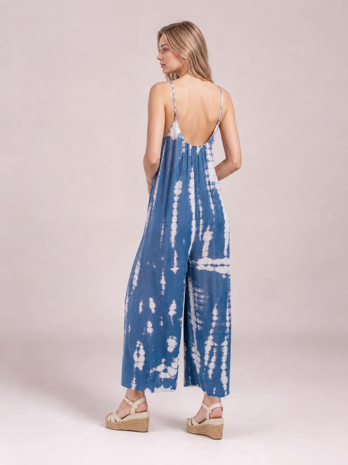 A model turns to show the back of a blue-and-white tie-dye spaghetti strap jumpsuit with a low back, relaxed wide-leg silhouette, and hands in pockets.