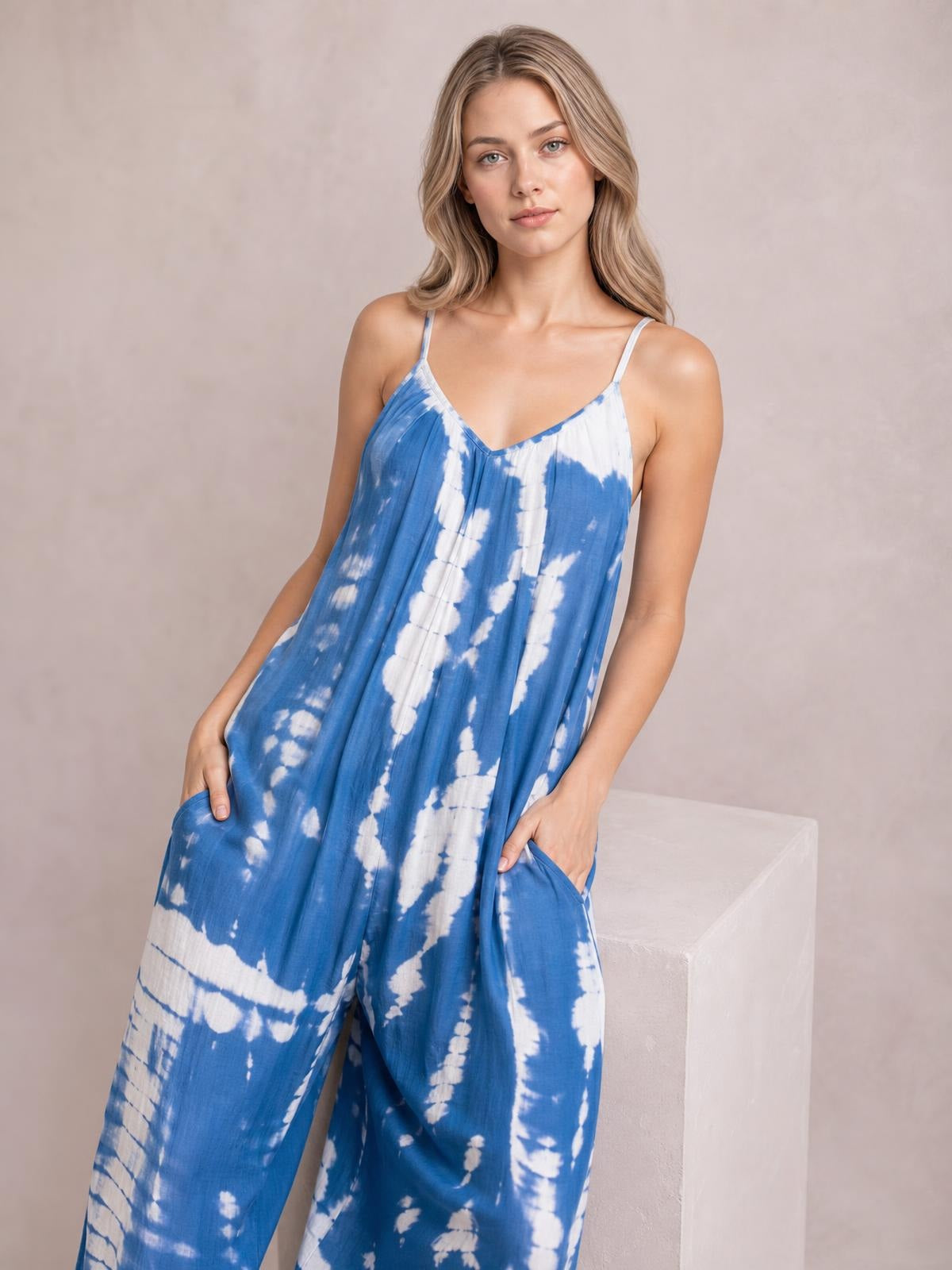 Model wears a blue and white tie-dye V-neck spaghetti strap jumpsuit with a relaxed wide-leg silhouette and hands tucked into its pockets.
