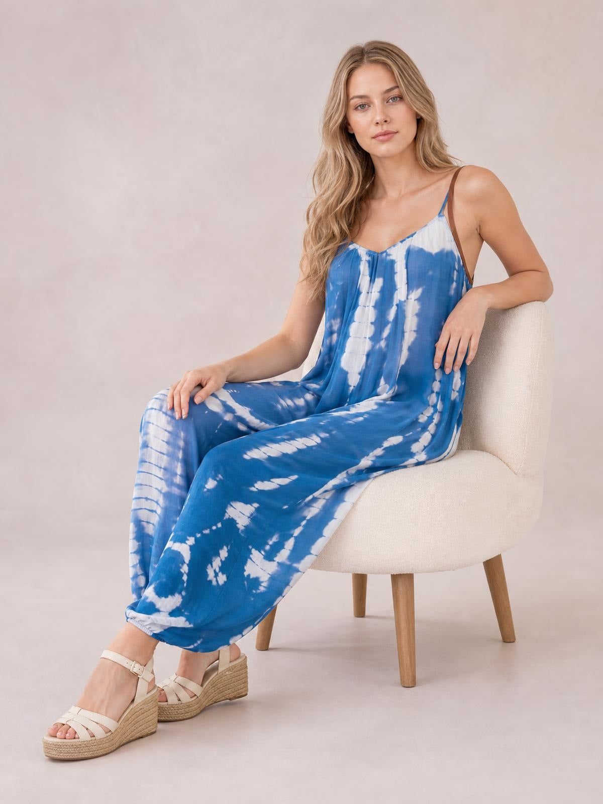 A seated model wears a blue-and-white tie-dye V-neck spaghetti strap jumpsuit with a flowing wide-leg silhouette, resting one arm on the chair.