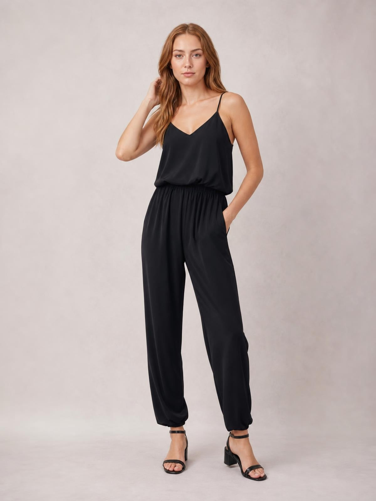 A model poses in a black V-neck spaghetti strap sleeveless jumpsuit with a smocked waistband, relaxed wide legs, and side pockets.