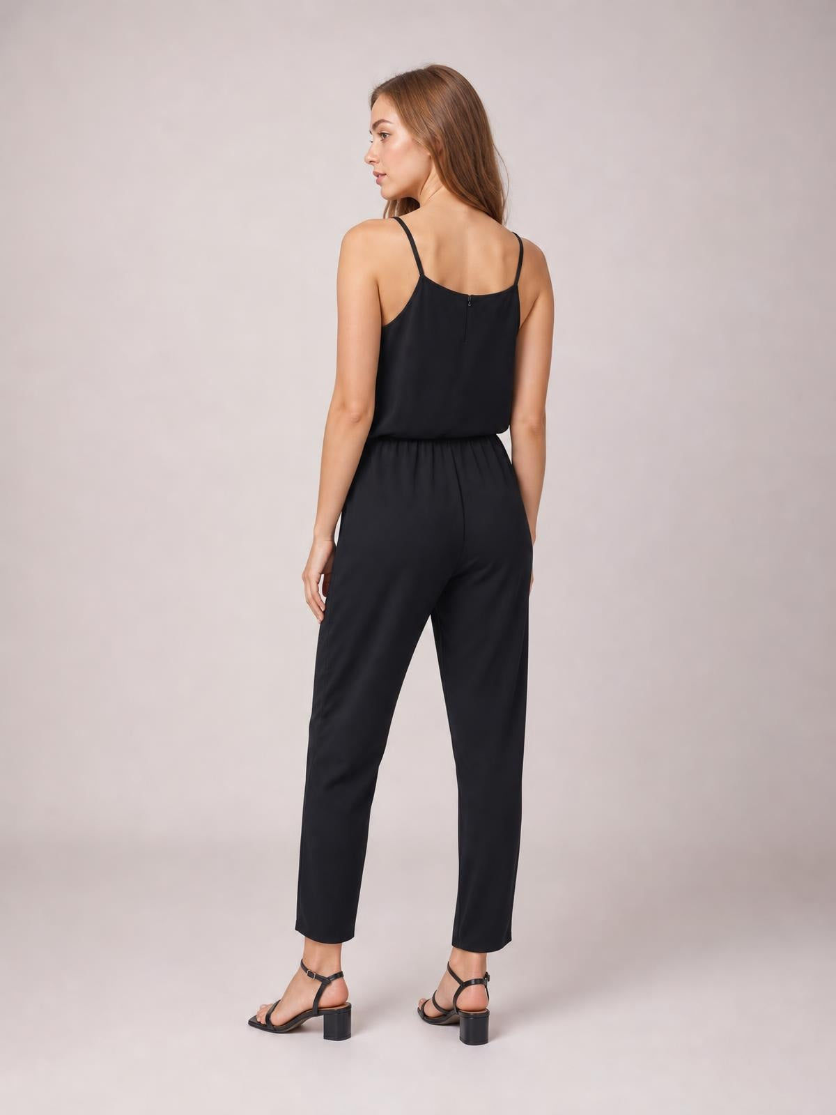 Model poses turned away in a black sleeveless jumpsuit with slim spaghetti straps, a gathered waist, and relaxed straight-leg silhouette.