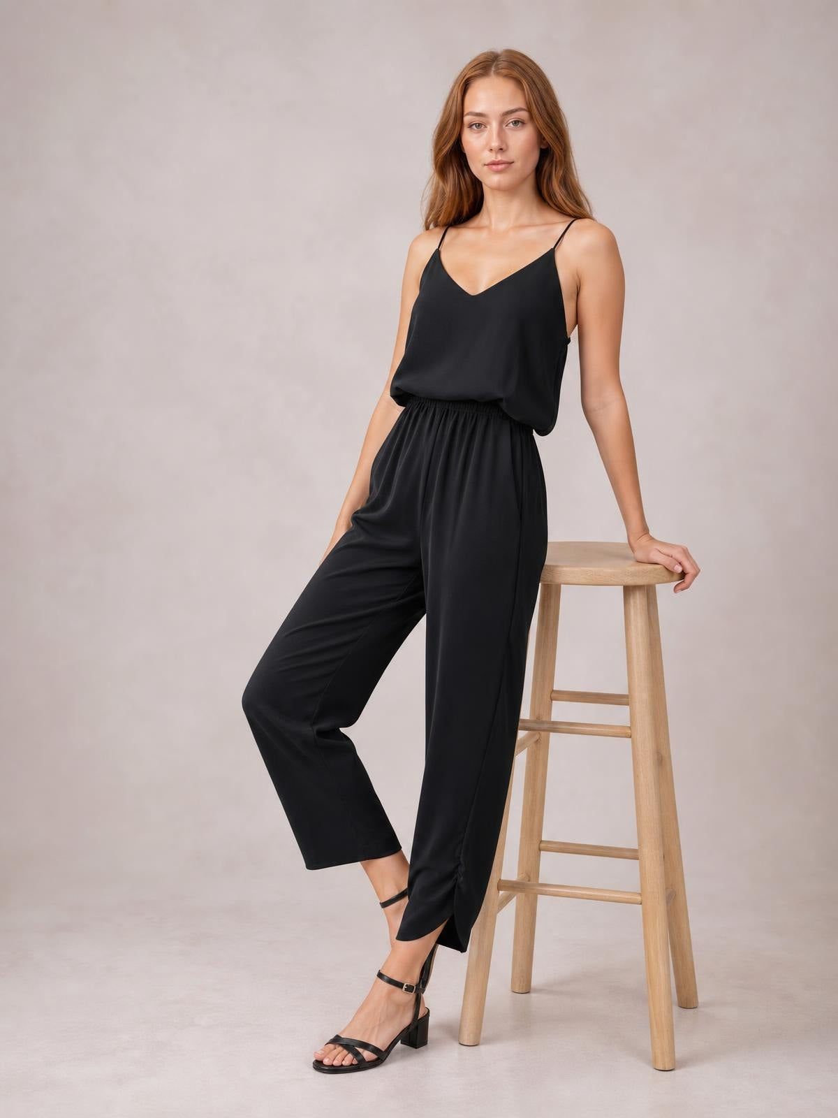 A model poses in a black V-neck spaghetti strap sleeveless jumpsuit with a smocked waist, wide-leg silhouette, and side pockets.