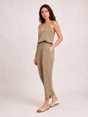 A model poses in a taupe V-neck spaghetti strap sleeveless jumpsuit with a smocked waist, relaxed tapered legs, and a hand tucked into its pocket.