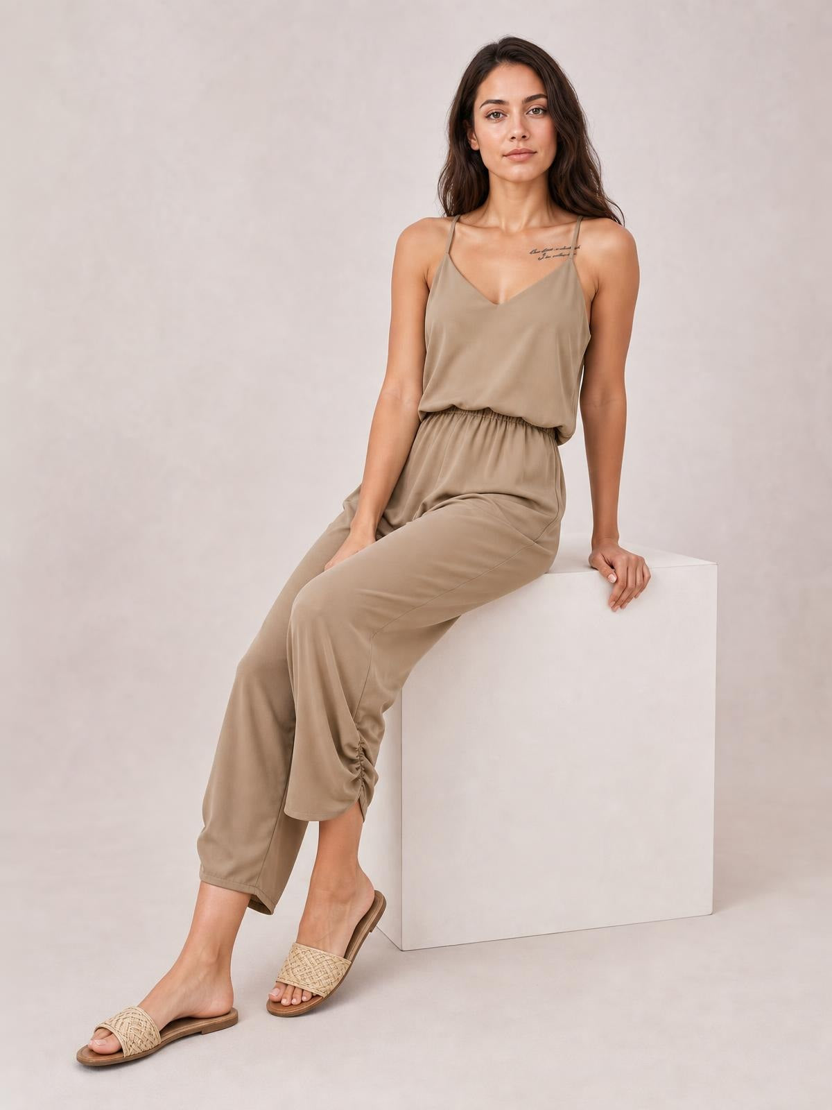 A model sits in a taupe V-neck spaghetti strap sleeveless jumpsuit with a smocked waist and relaxed wide-leg silhouette.
