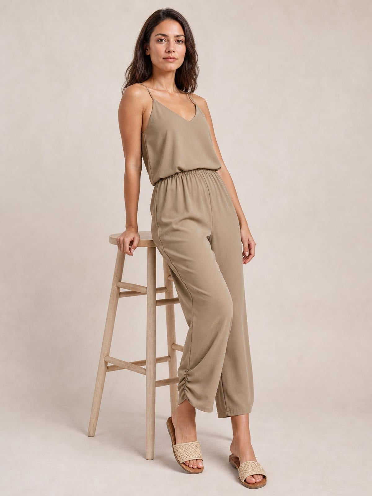 A model leans on a stool wearing a taupe V-neck spaghetti strap sleeveless jumpsuit with a smocked waist and relaxed cropped wide legs.