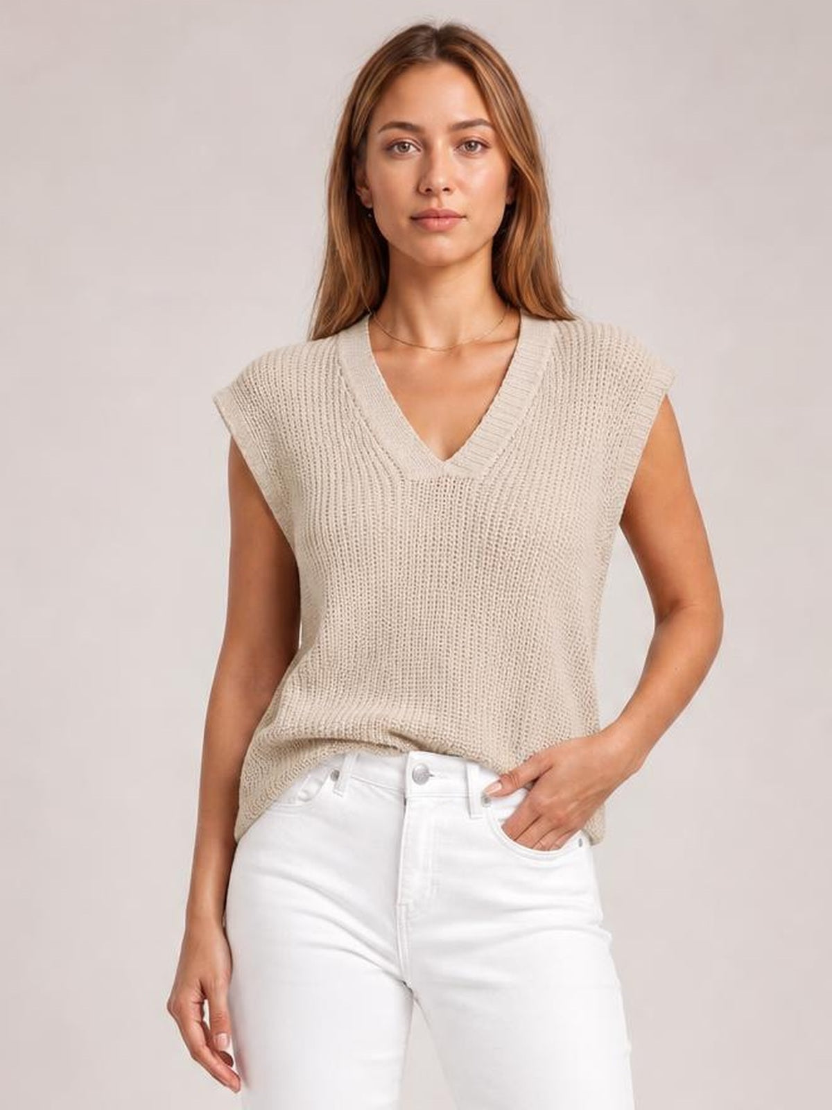 A woman wears a cream V-neck sweater vest with ribbed trim, sleeveless construction, textured knit, and a relaxed straight silhouette over white jeans.
