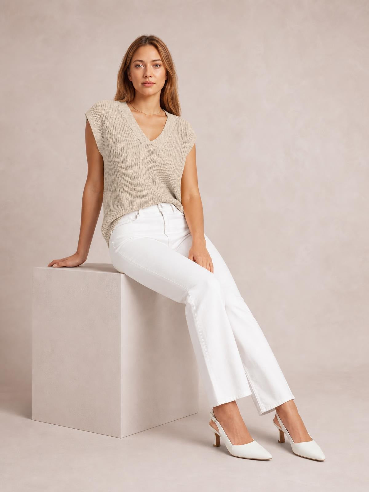 Model seated in a beige V-neck sweater vest with a textured knit, ribbed edges, sleeveless cut, and relaxed straight silhouette.