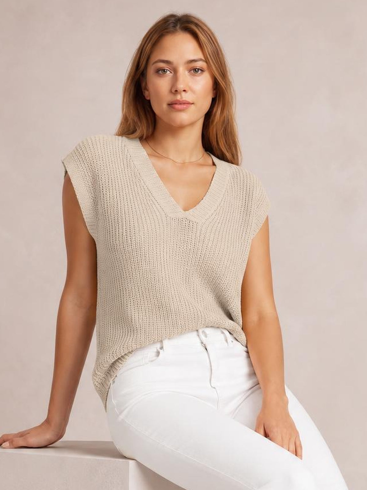 Model seated and leaning on one hand wears a light beige V-neck sweater vest with a textured knit, relaxed straight silhouette, and wide sleeveless shoulders.