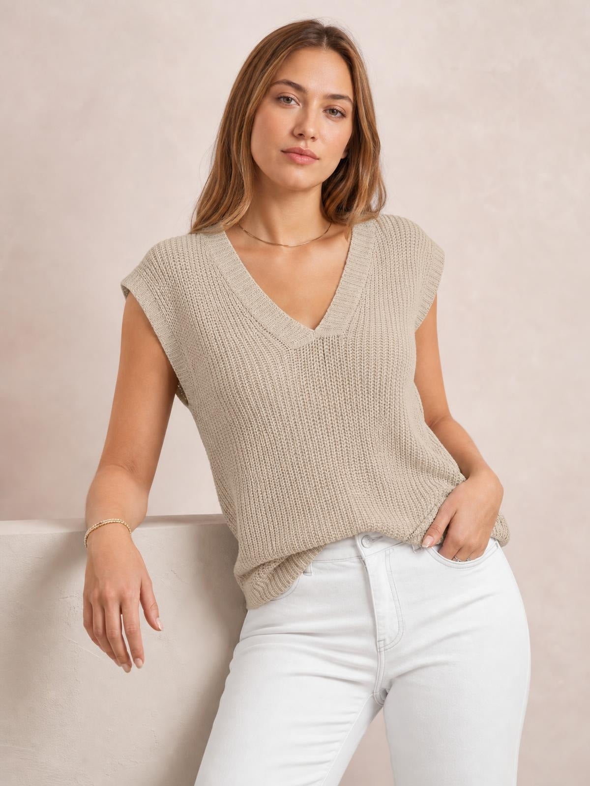 Model wears a beige V-neck sweater vest with ribbed knit texture, sleeveless cut, and relaxed straight silhouette while leaning with one hand in her pocket.