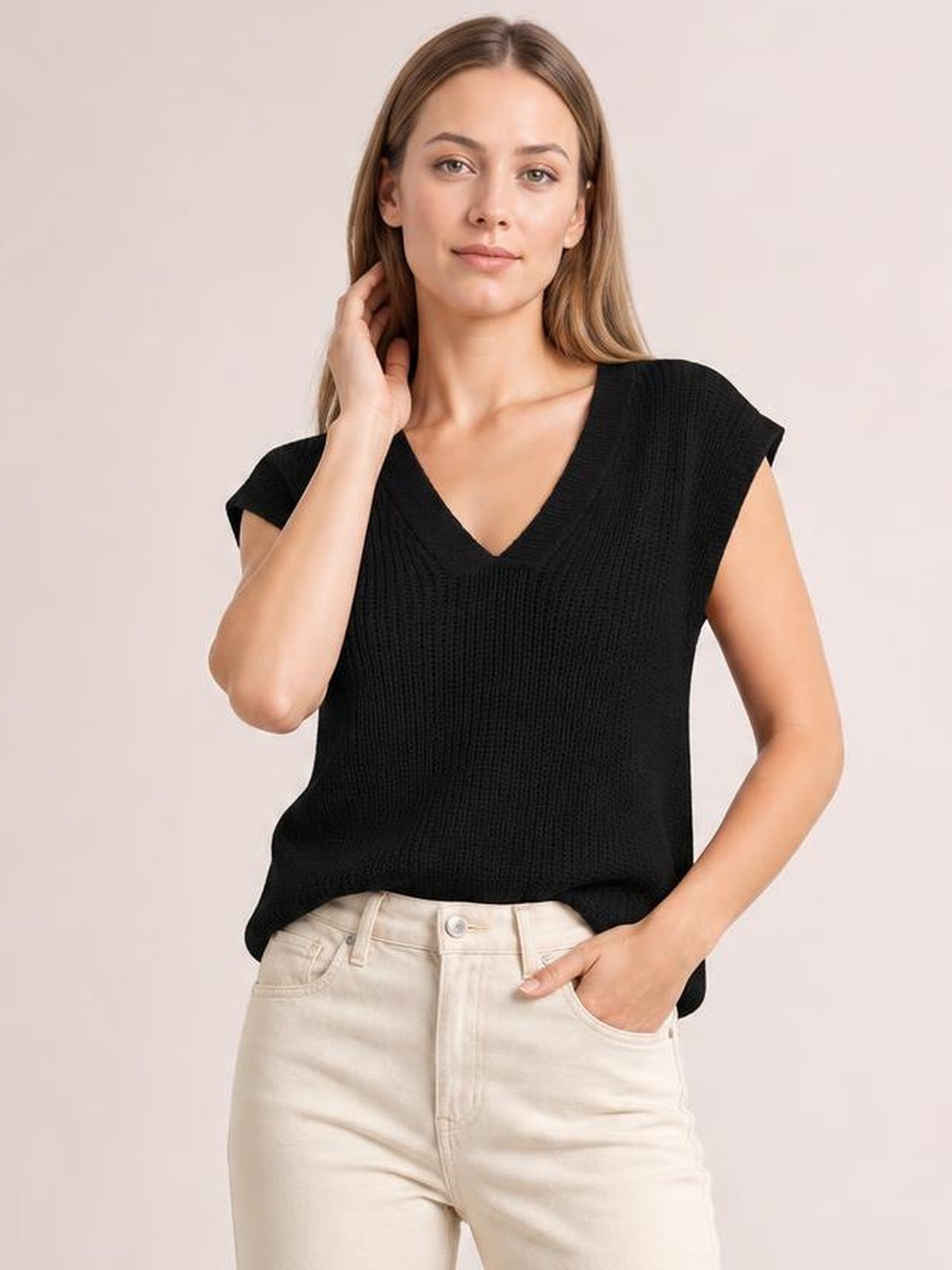Model wearing a black V-neck sweater vest with a textured knit, cap sleeves, relaxed silhouette, and slightly curved hem, posing with one hand in her pocket.