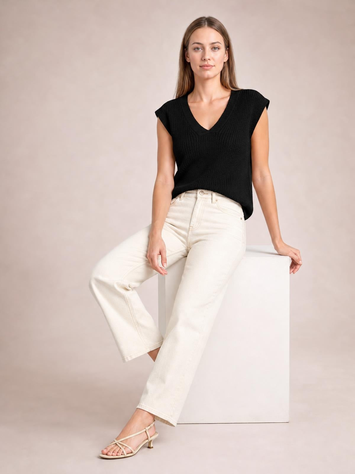 Model seated in a relaxed pose wearing a black V-neck sweater vest with ribbed knit texture, cap sleeves, and a loose straight silhouette.