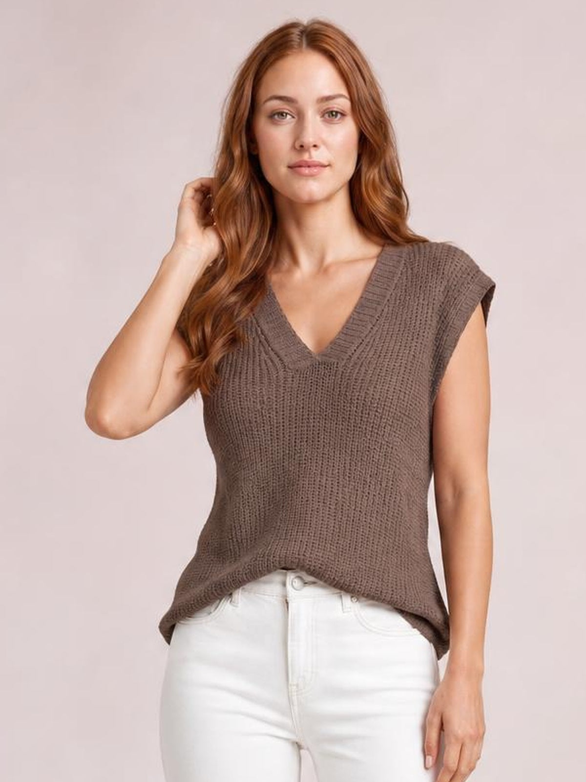 Model poses in a brown knitted V-neck sweater vest with ribbed trim, cap sleeves, a relaxed straight silhouette, and curved hem over white pants.