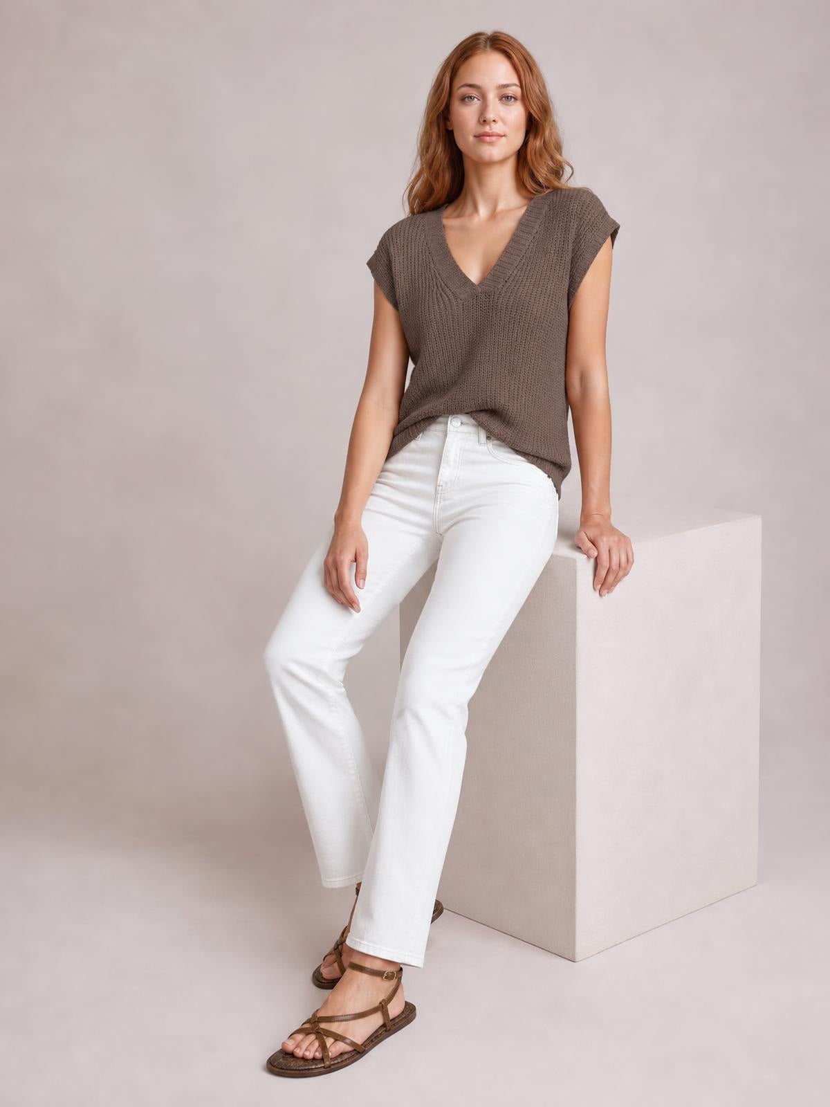 Model wearing a taupe V-neck sweater vest with a chunky ribbed knit, wide shoulder openings, and a relaxed straight silhouette while seated.