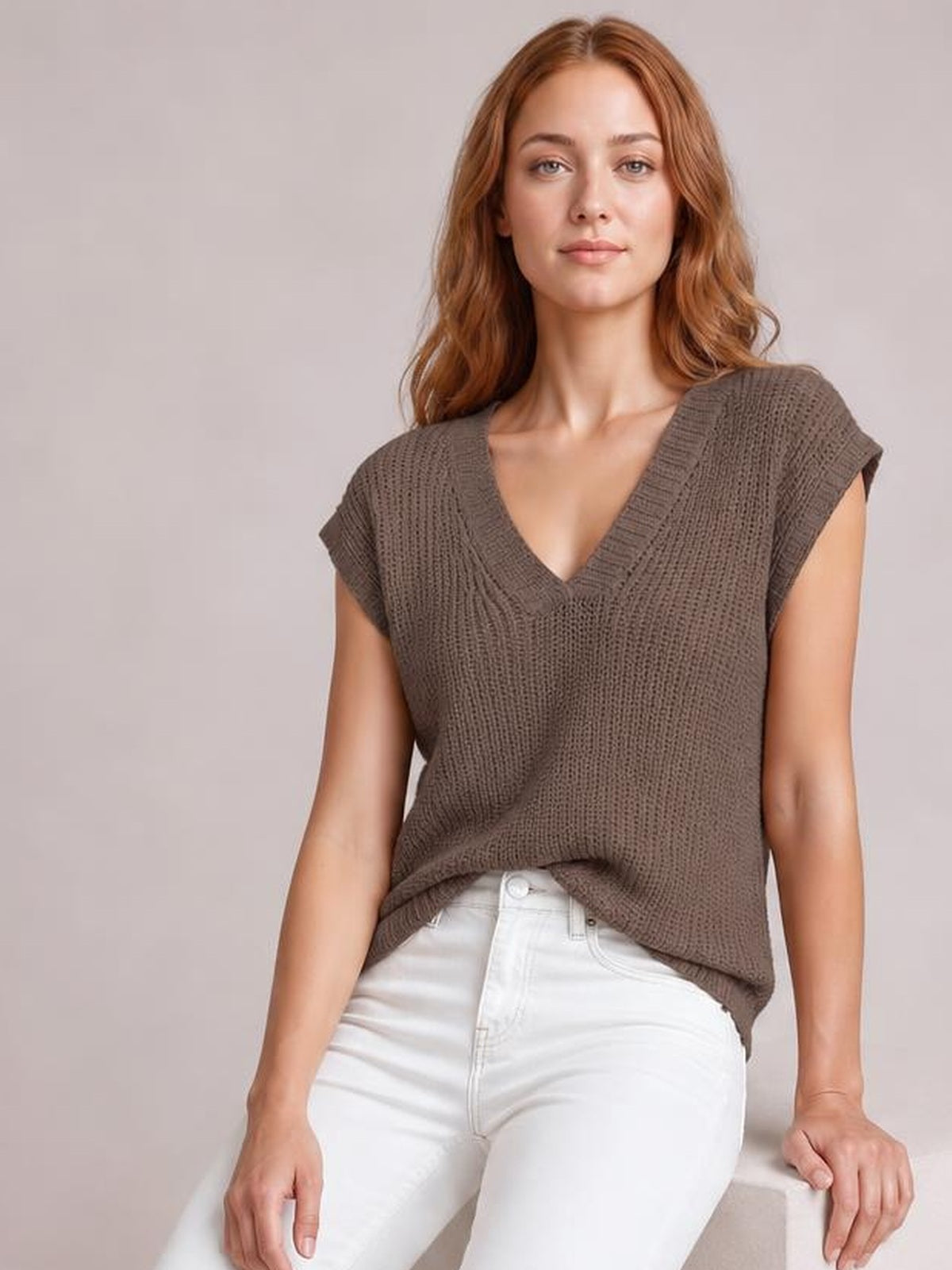 Model wears a taupe V-neck sweater vest with textured knit construction, ribbed trim, cap sleeves, and a relaxed silhouette while seated.