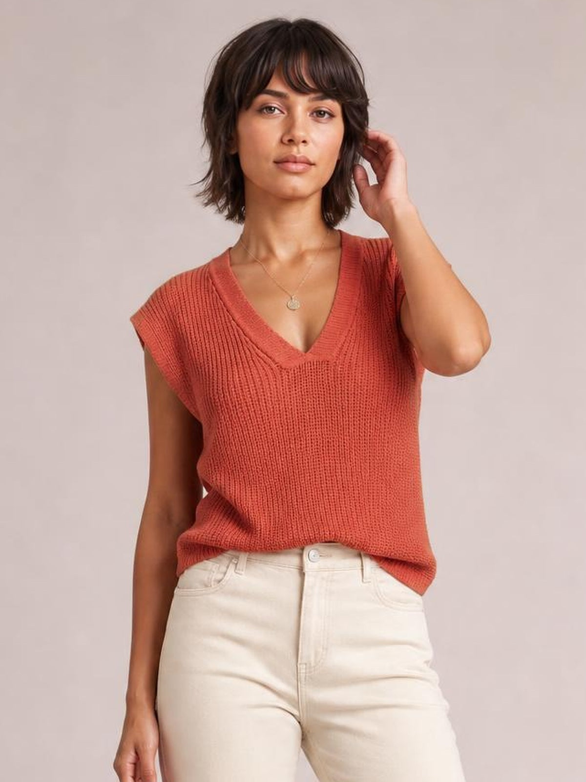 Model poses in a coral V-neck sweater vest with ribbed knit texture, wide shoulder openings, and a relaxed silhouette worn with light-colored jeans.