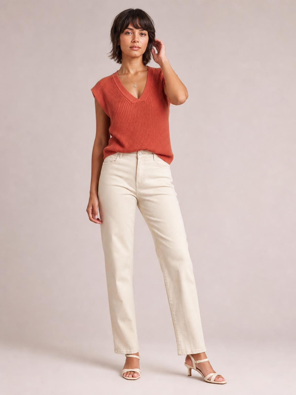 Model wears a coral V-neck sweater vest with ribbed knit construction, relaxed sleeveless silhouette, and a slightly draped hem while posing with one hand raised.