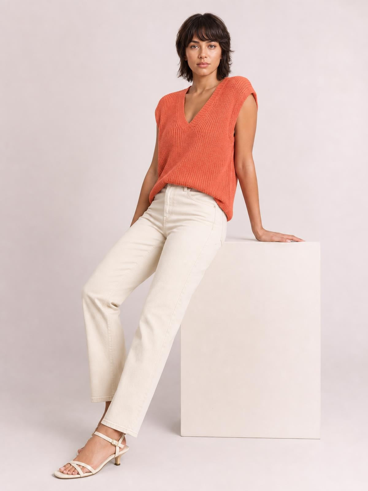 Model wears a coral V-neck sweater vest with a ribbed knit texture, sleeveless cut, relaxed silhouette, and wide ribbed neckline and armholes.