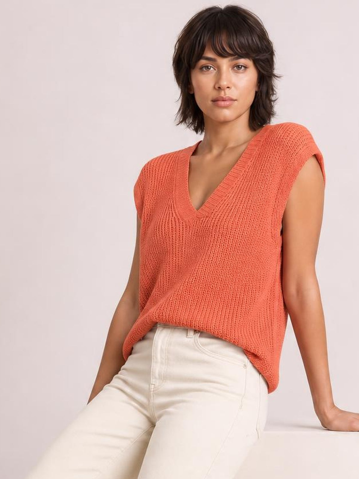 Model wearing a coral V-neck sweater vest with a relaxed knit silhouette, ribbed neckline, sleeveless construction, and slightly stretchy texture.