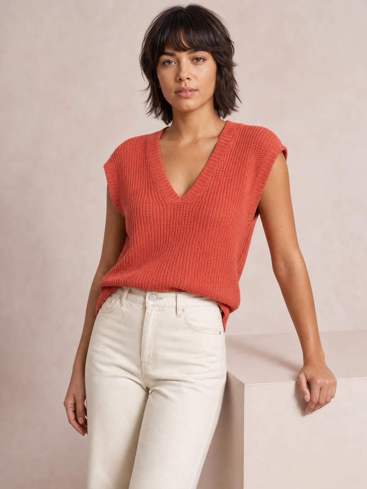 Model wearing a coral V-neck sweater vest with ribbed knit texture, sleeveless silhouette, and wide bound edges, styled with cream jeans.