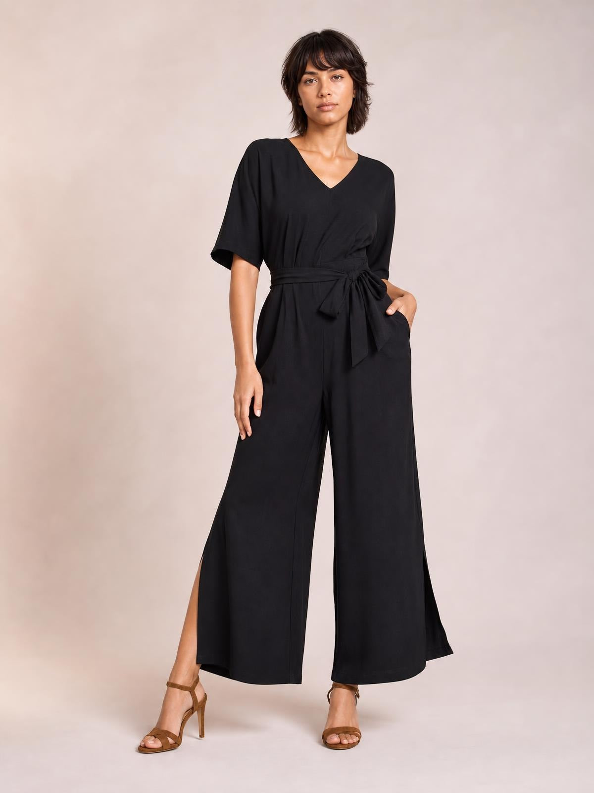 Model poses in a black V-neck jumpsuit with short sleeves, a tied waist, wide-leg silhouette, and side slits at the hems.