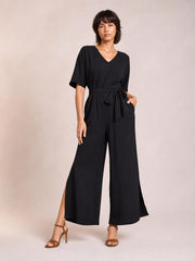 Model poses in a black V-neck jumpsuit with short sleeves, a tied waist, wide-leg silhouette, and side slits at the hems.