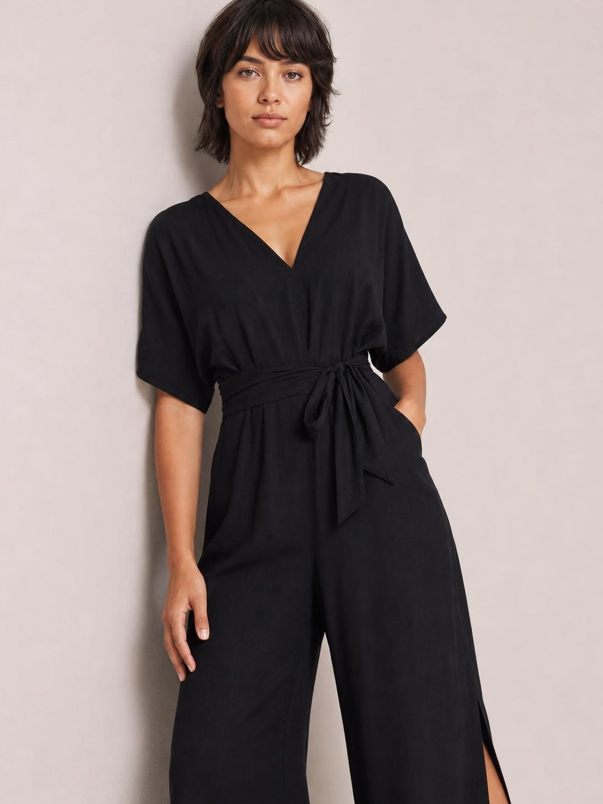 Model wears a black V-neck jumpsuit with wide-leg silhouette, short sleeves, a tied waist detail, and a visible side slit.