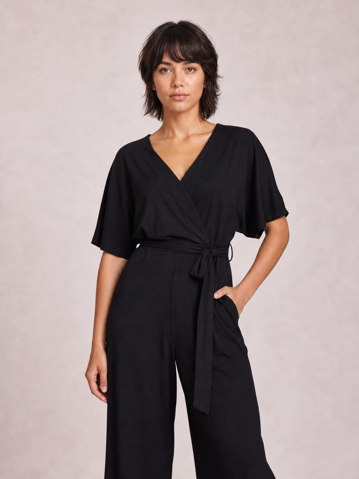 A model stands in a black V-neck jumpsuit with short sleeves, a tied waist, flowing wide-leg silhouette, and one hand resting in a pocket.