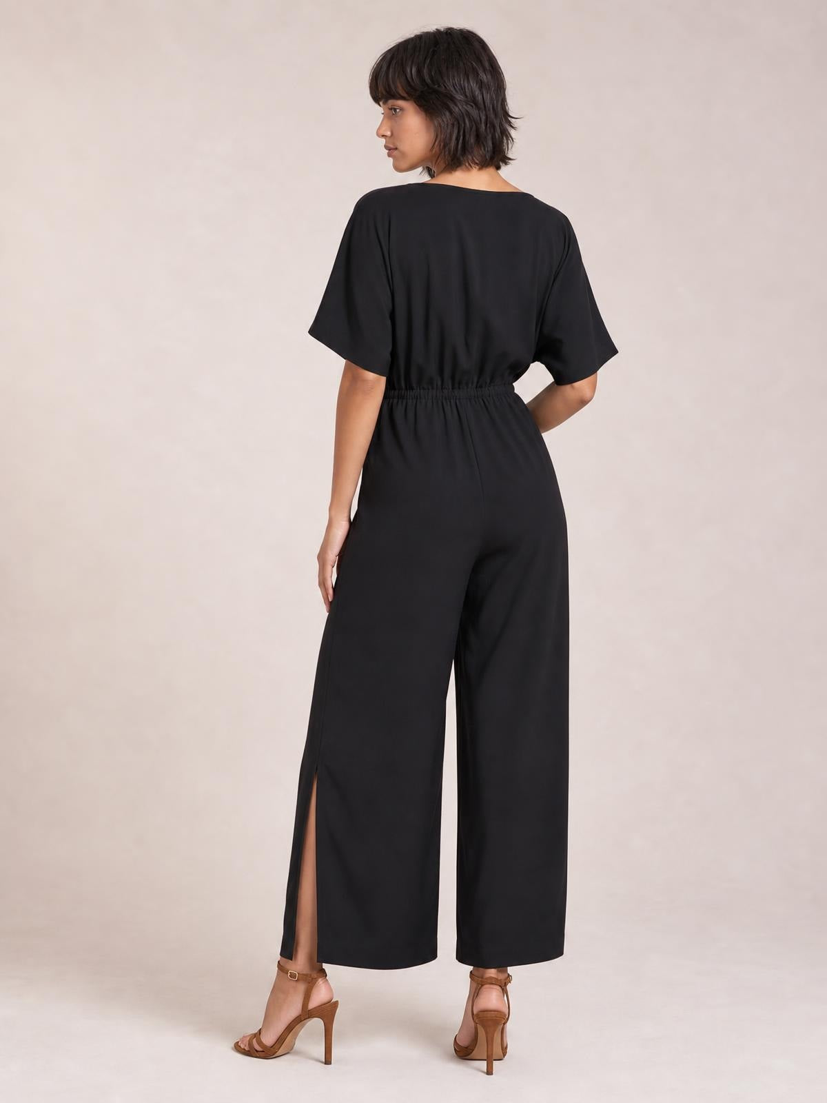 A model poses in a black jumpsuit with short sleeves, a gathered waist, wide-leg silhouette, and a visible side slit at the ankle.