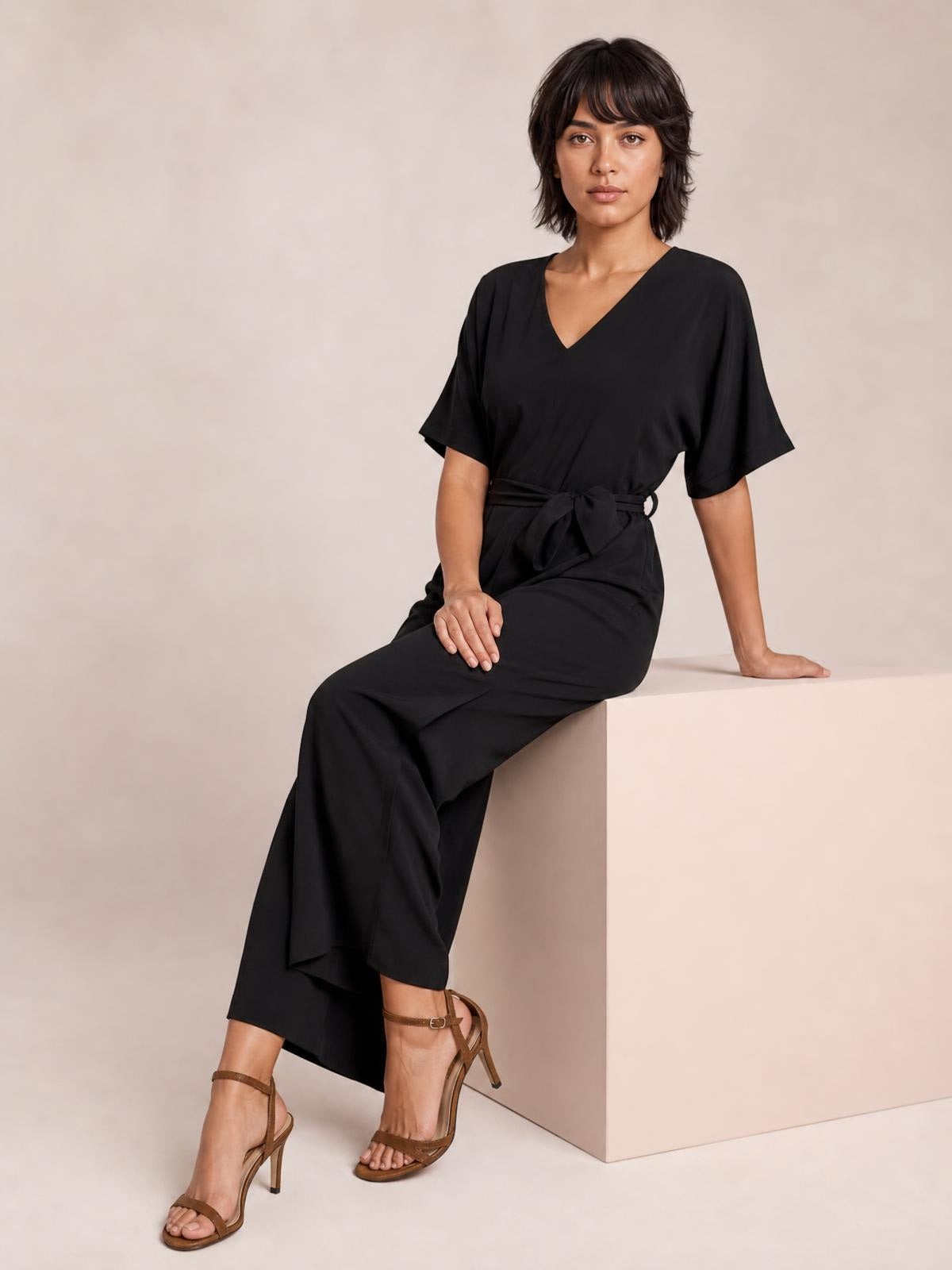 Model seated in a black V-neck jumpsuit with short sleeves, a tied waist, wide-leg silhouette, and flowing opaque fabric.