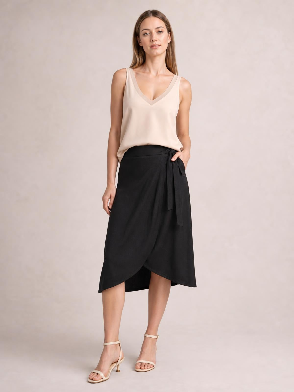 Model wears a beige V-neck wide strap cami with a relaxed, fluid silhouette, standing with one hand in the pocket of a black wrap skirt.