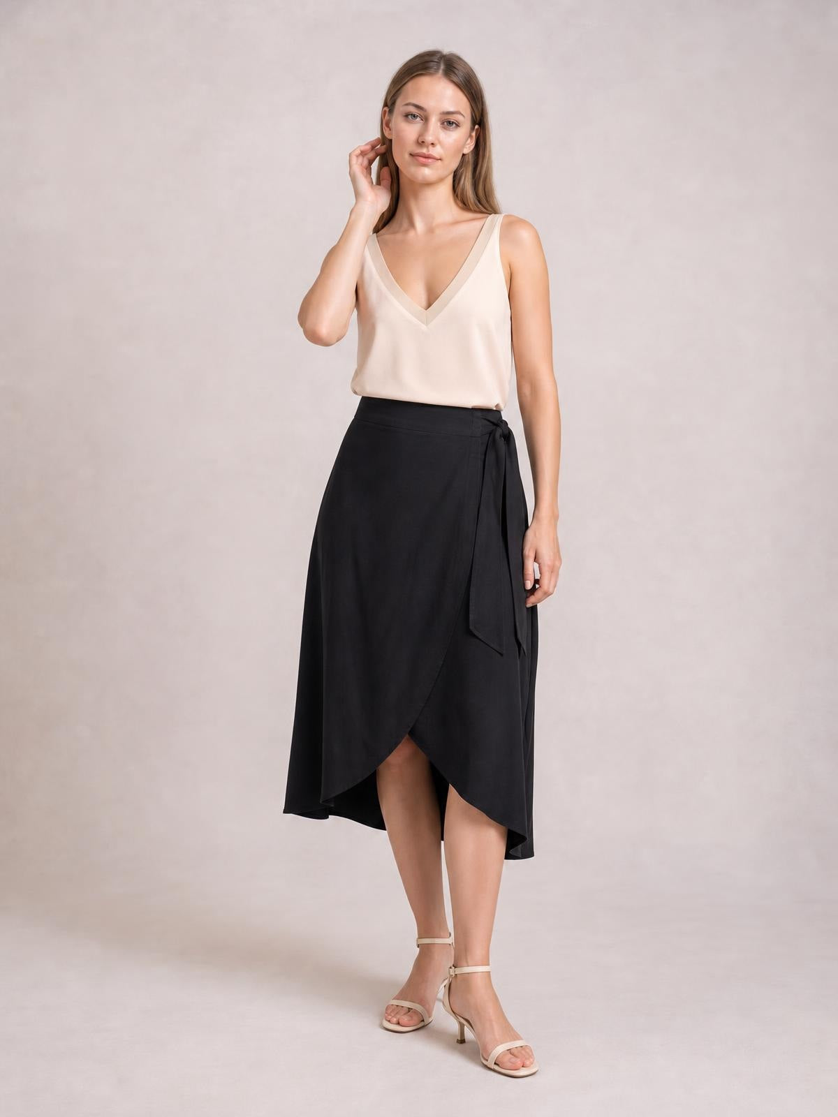 Model wearing a cream V-neck wide strap cami with a relaxed silhouette, smooth opaque fabric, and clean construction, posing with one hand near her hair.
