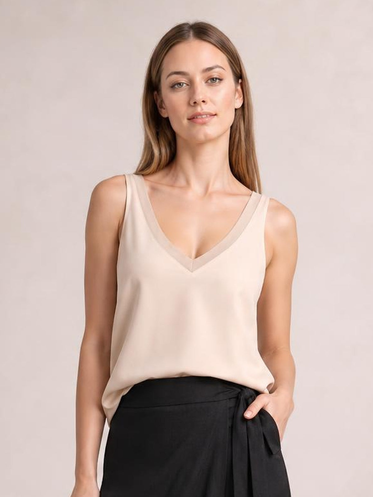 Model wears a cream V-neck wide strap cami with a softly draped, relaxed silhouette and subtle neckline trim, standing with one hand in her pocket.