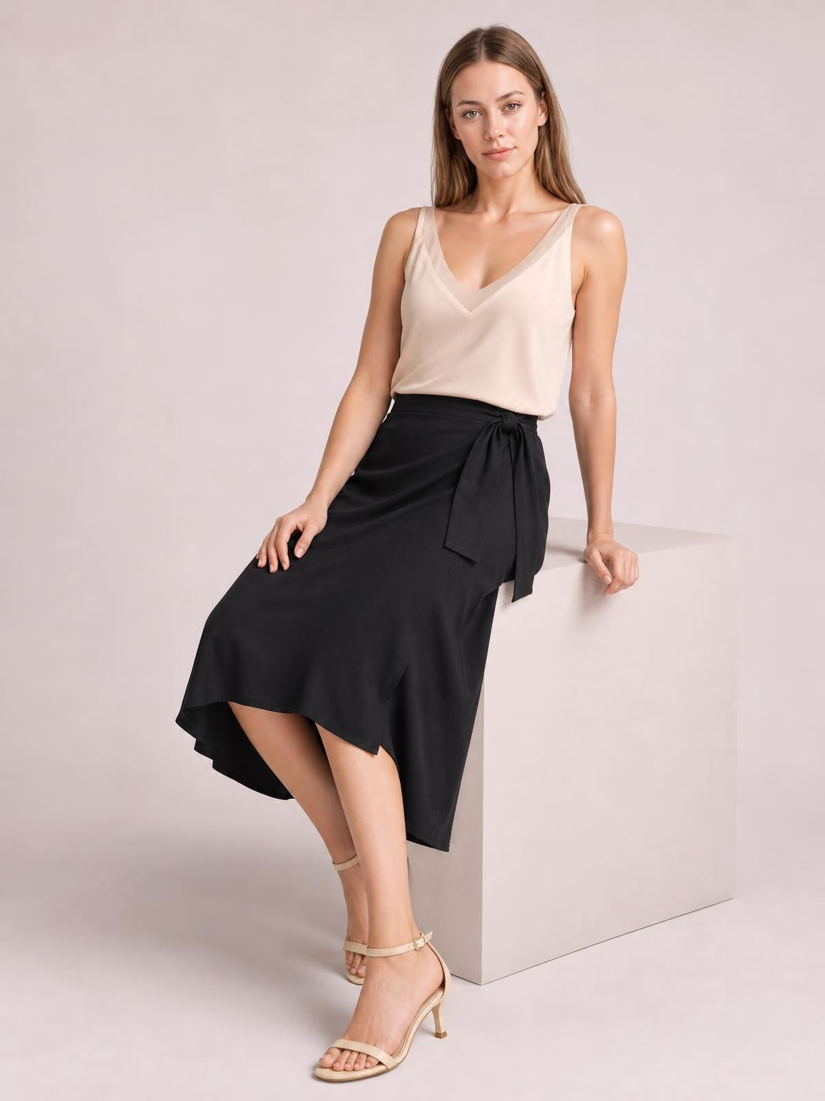 A model wears an opaque blush-beige V-neck wide strap cami with a relaxed, fluid silhouette while seated with one hand resting beside her.