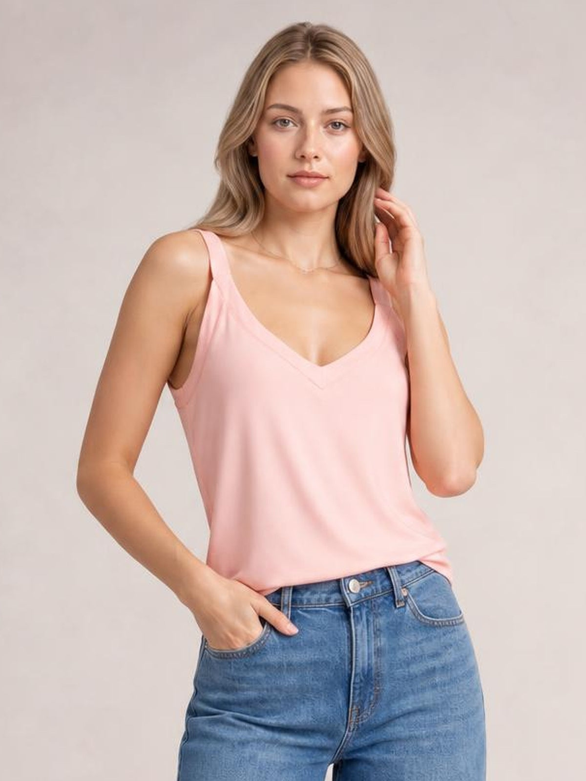 A model wears a pink V-neck wide strap cami with a relaxed straight silhouette, posing with one hand in her jeans pocket and the other touching her hair.