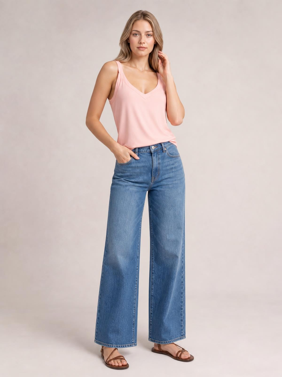 Model wearing a pink V-neck wide strap cami with a relaxed, straight silhouette, posed with one hand in her jeans pocket and the other touching her hair.