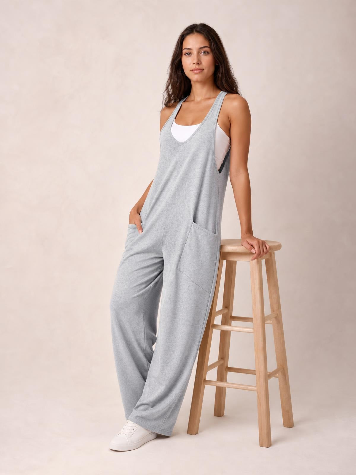 A model leans against a stool in gray V-neck wide-strap overalls with a relaxed wide-leg silhouette, deep side pockets, and a white tank underneath.