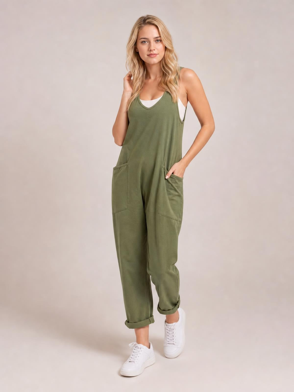 A model poses in olive-green V-neck wide-strap overalls with a relaxed cropped-leg silhouette, cuffed hems, and large front patch pockets over a white tank.