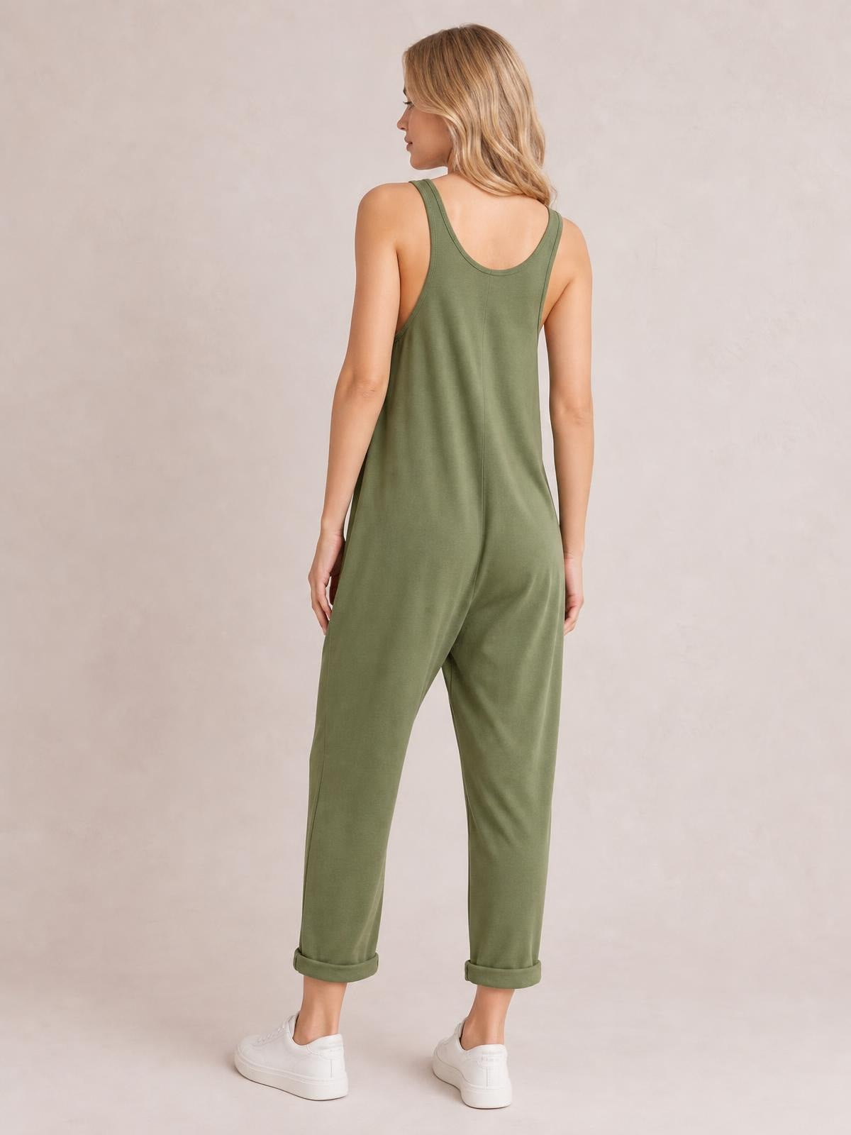 A model stands turned away in olive-green wide-strap overalls with a relaxed, straight-leg silhouette, center-back seam, scoop back, and rolled ankle cuffs.