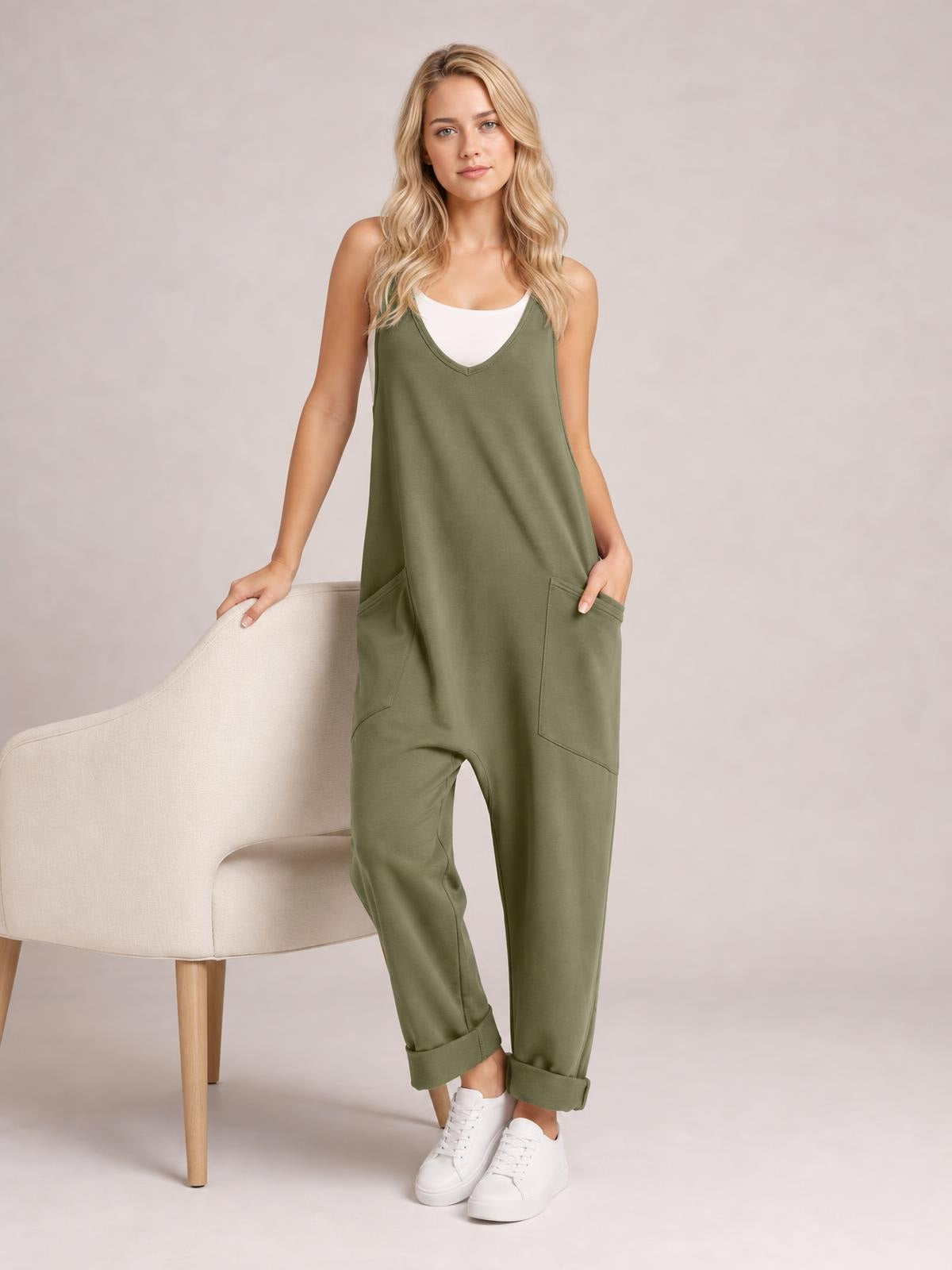 A model poses in olive-green V-neck wide-strap overalls with a relaxed jumper silhouette, roomy front pockets, and rolled cuffs, one hand tucked inside.