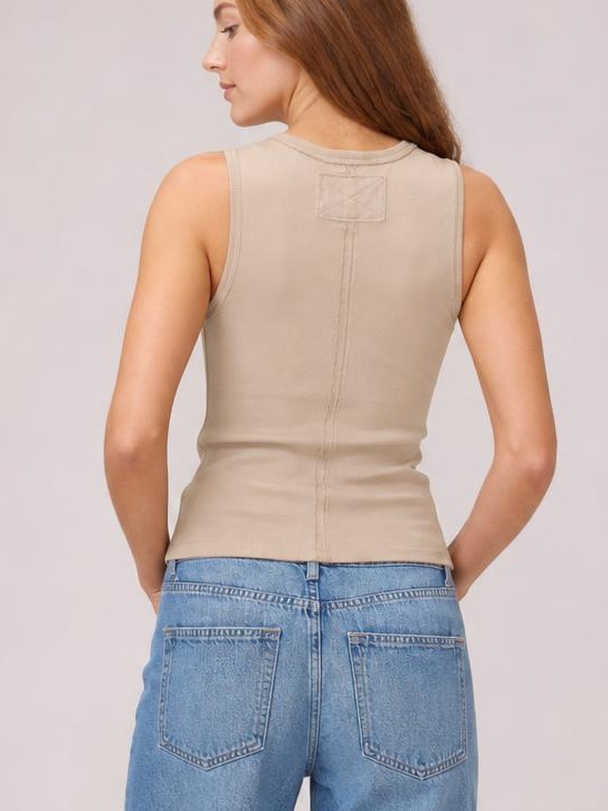 A model faces away in a beige wide-strap tank with a fitted silhouette, high back neckline, central seam, and visible upper-back patch.