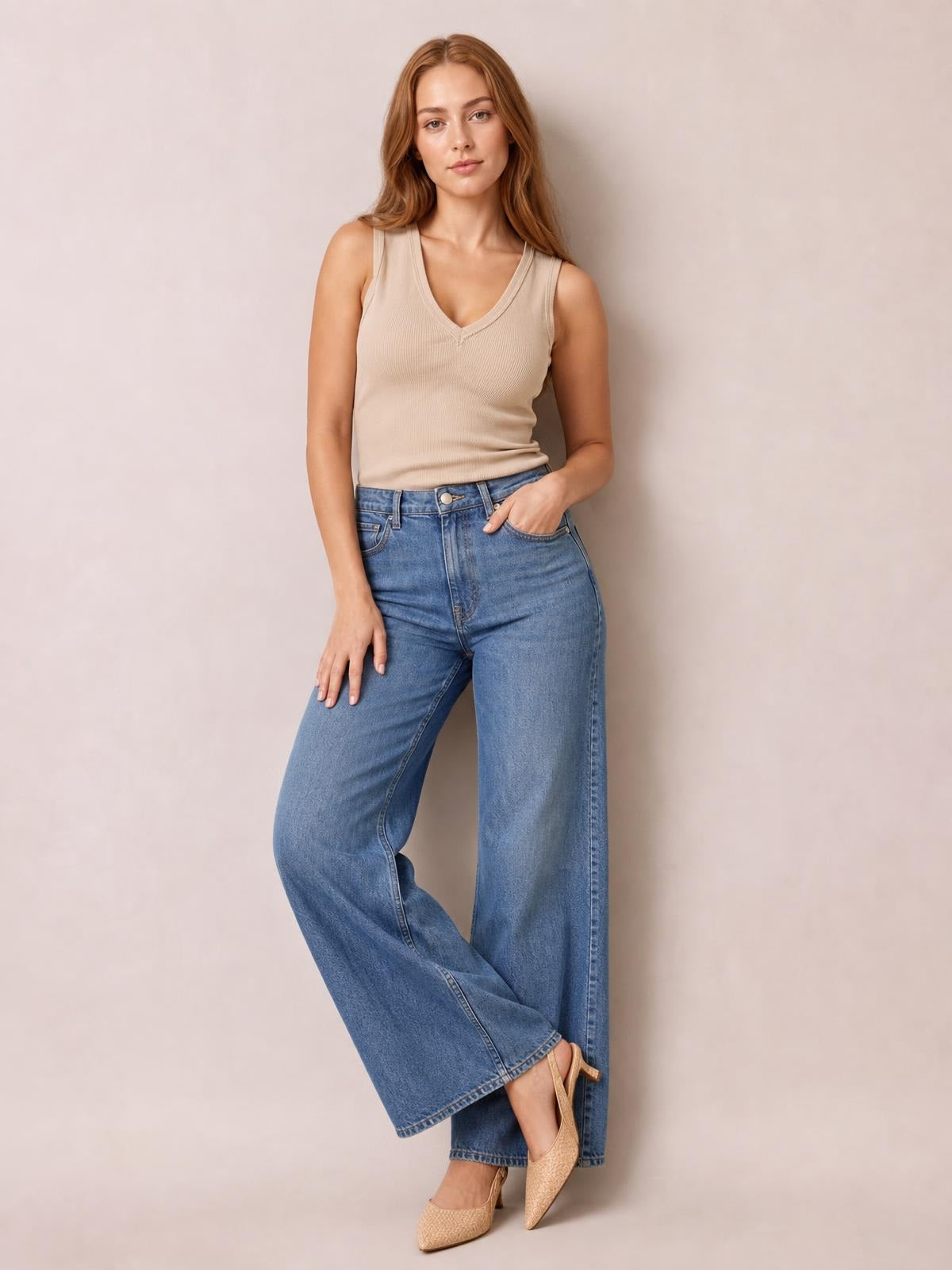 A model poses in a fitted beige V-neck wide strap tank with subtle ribbing, paired with wide-leg jeans and one hand resting in a pocket.