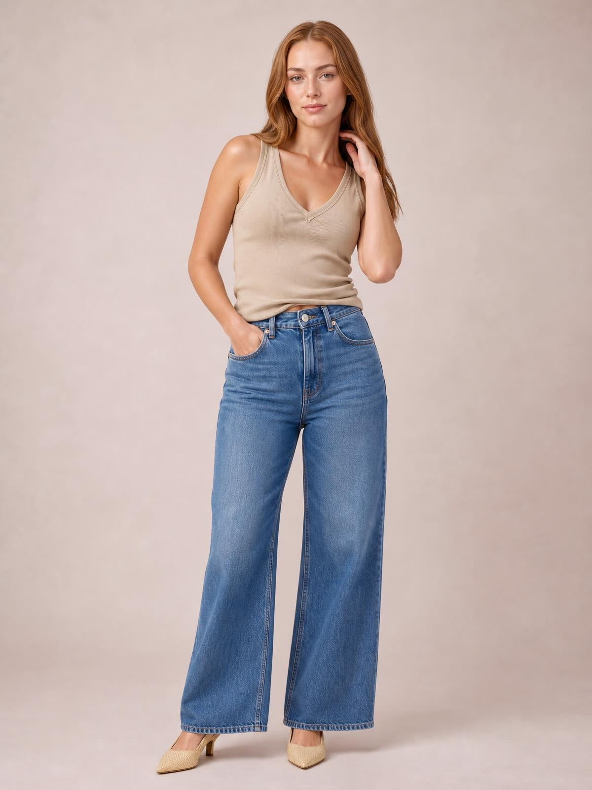 A model poses in a beige V-neck wide strap tank with a close-fitting silhouette and subtle ribbed texture, one hand in her pocket and the other touching her hair.