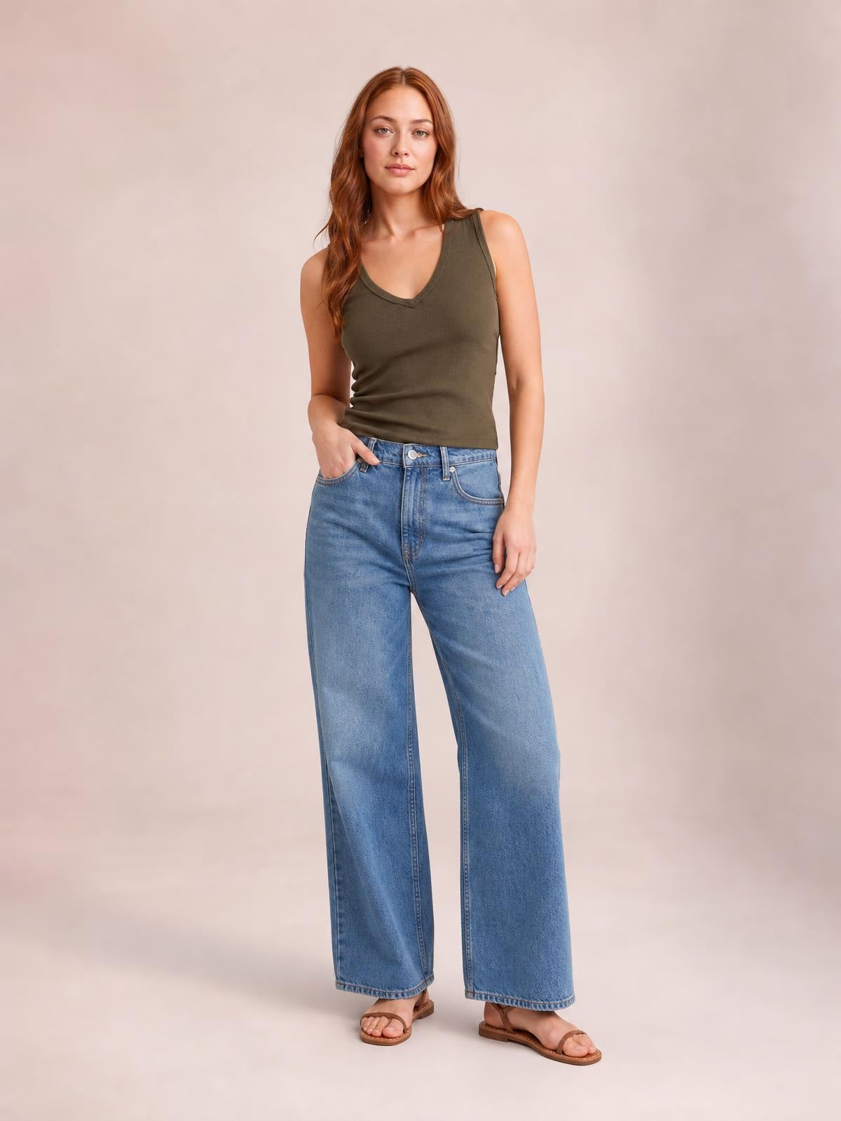 Model wearing an olive green V-neck wide strap tank with a fitted, subtly stretchy silhouette and ribbed construction, paired with wide-leg jeans.
