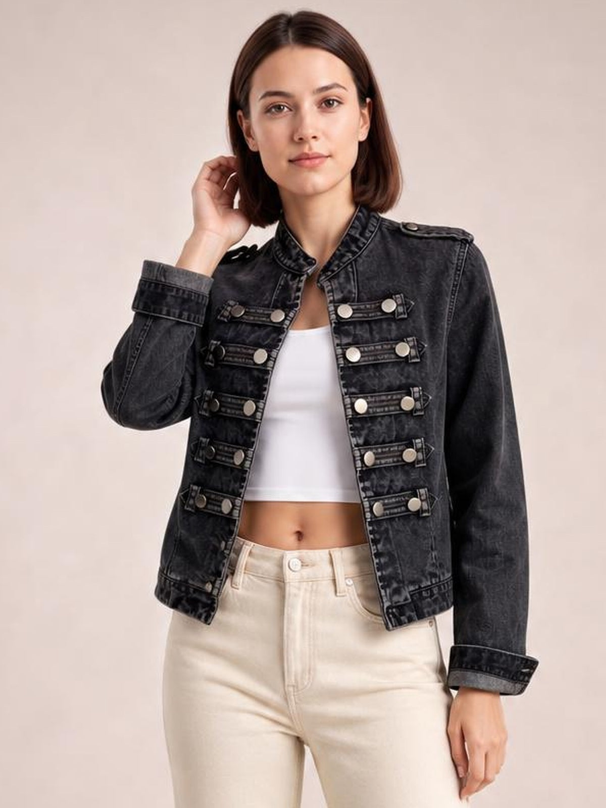 Model poses with one hand near her hair wearing a dark washed denim jacket with an open front, stand collar, long sleeves, cropped silhouette, and rows of decorative buttoned bands.