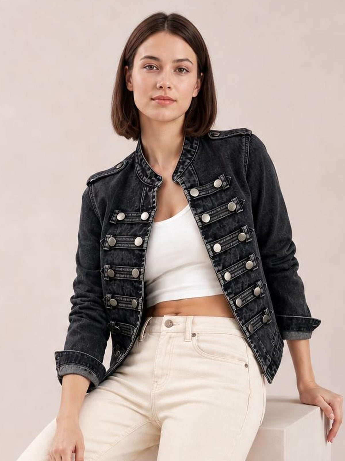 Model seated in a washed black denim jacket with an open front, decorative buttoned chest details, structured collar, shoulder tabs, and rolled sleeves.