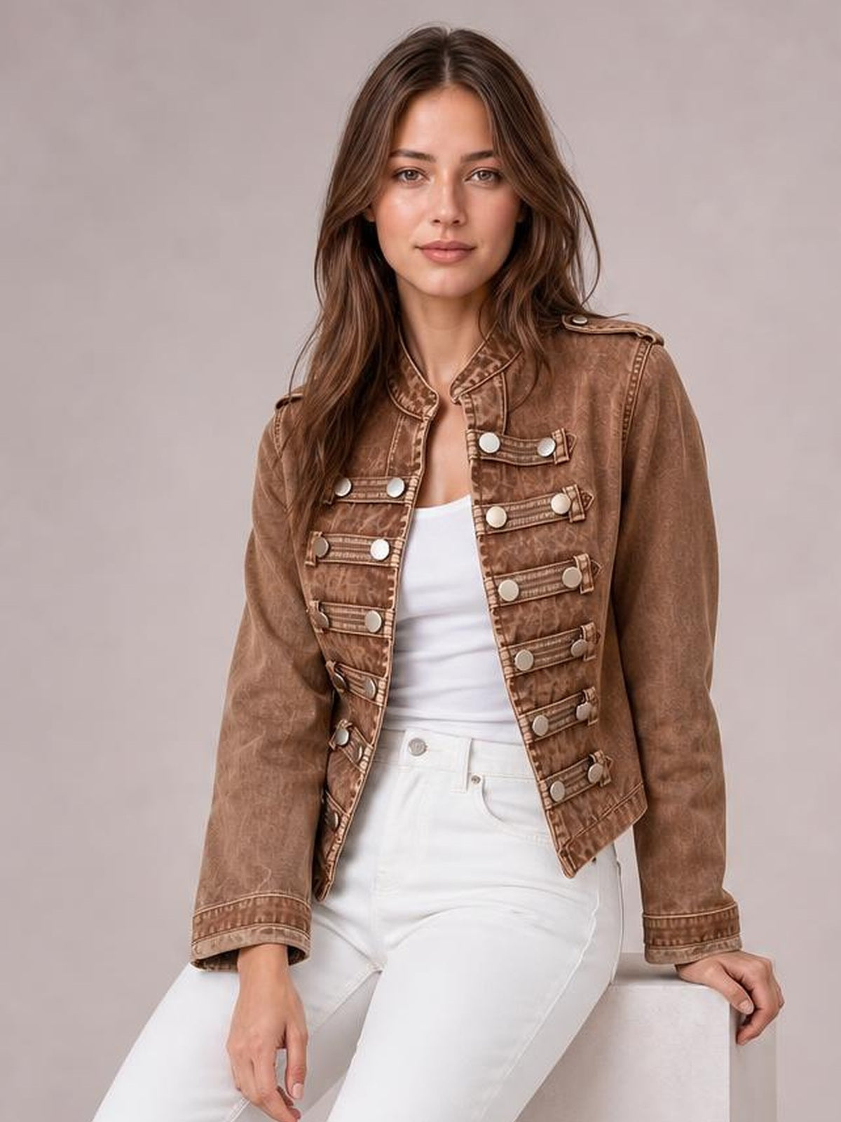 A model wears a brown washed denim jacket with an open front, long sleeves, shoulder tabs, and rows of decorative buttoned straps in a fitted silhouette.