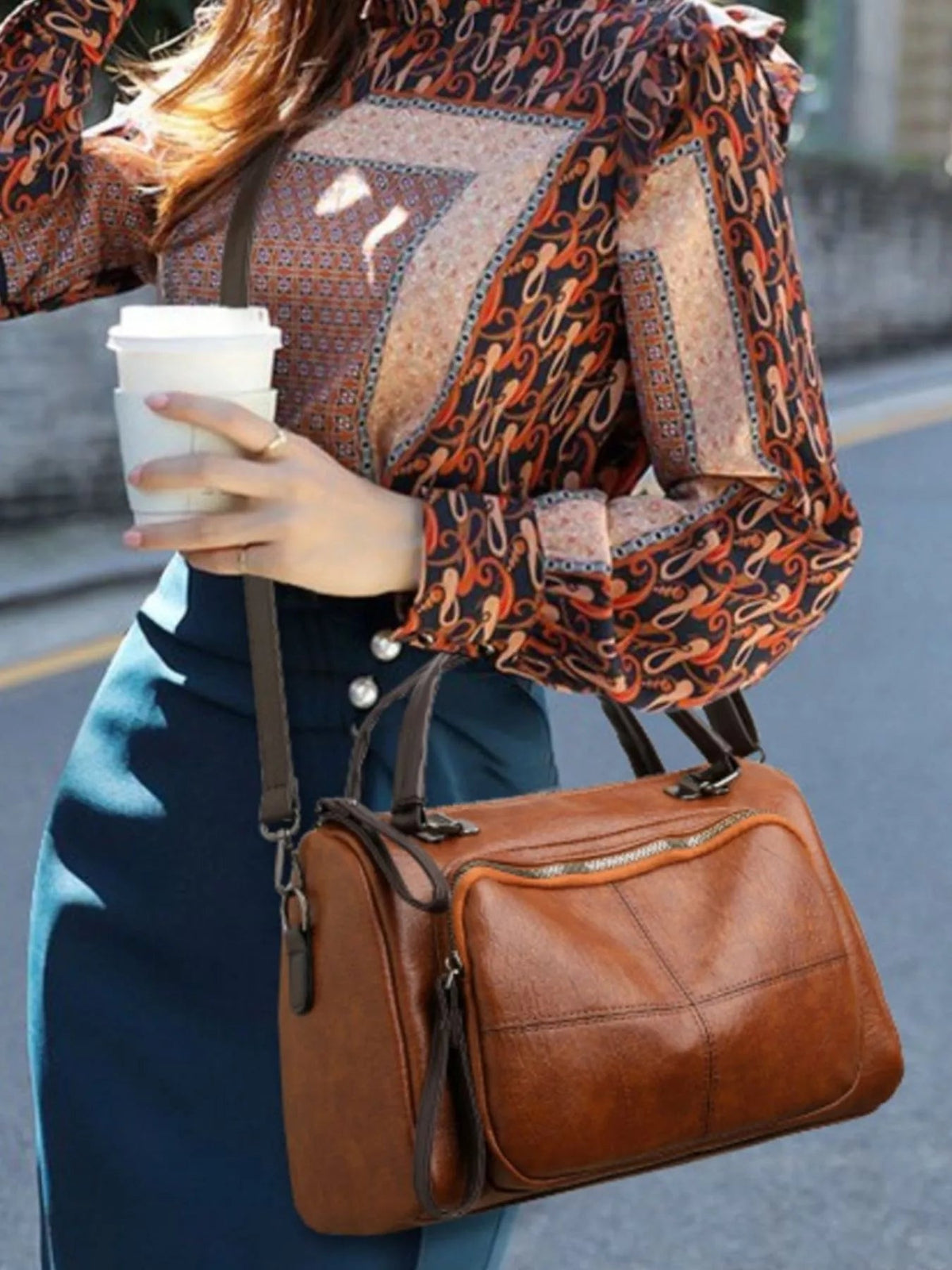 A model in a patterned blouse carries a medium vintage-style brown PU leather crossbody bag with rounded edges, a front zip pocket, and a long strap.