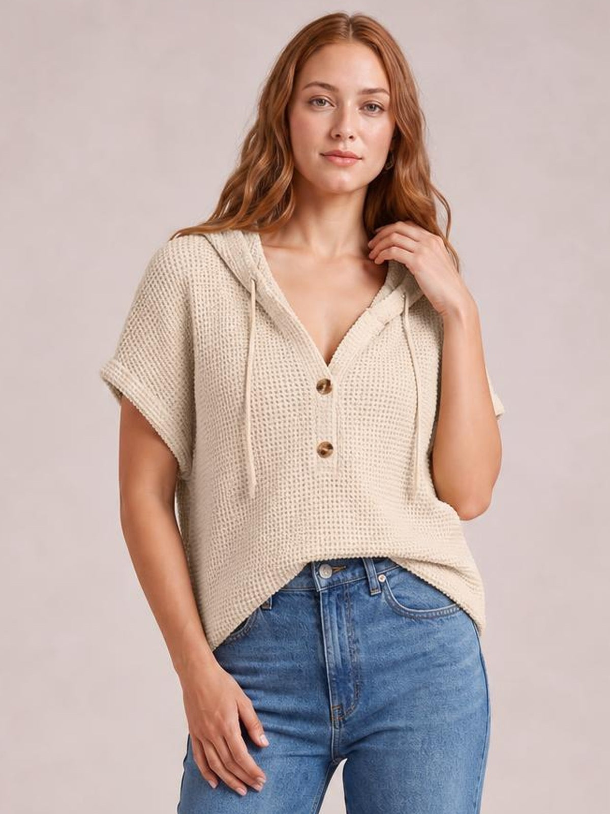 Model wears a cream waffle-knit hooded short-sleeve top with a relaxed, slightly cropped silhouette, drawstring hood, and two-button front closure.